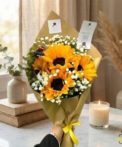 Hand-held sunflower bouquet wrapped in brown paper with baby’s breath flowers, placed in a cozy indoor setting.